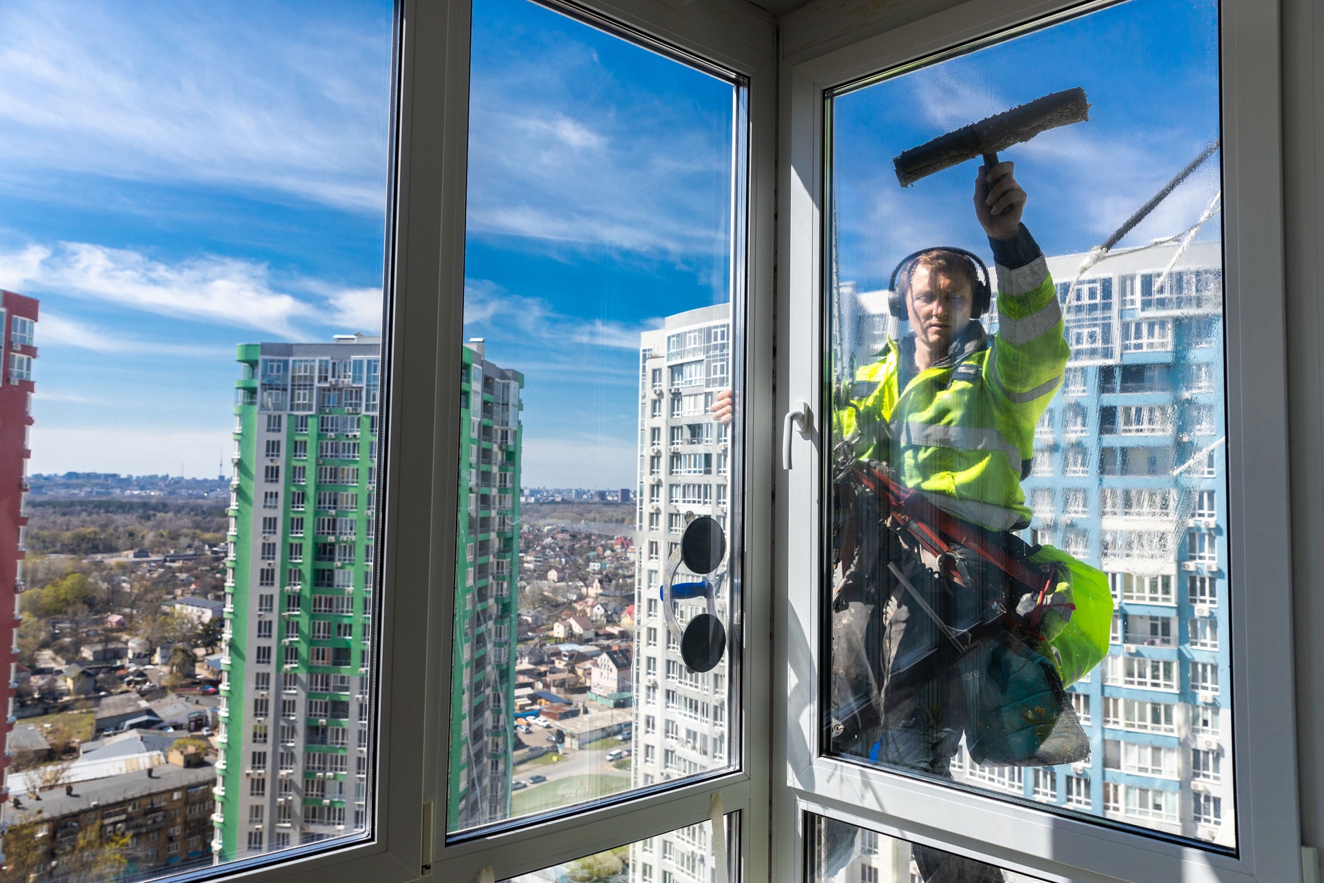 Professional Window Cleaner Working at Heights in an Urban Setting with a Scenic View of High-Rise Buildings in the Background and a Clear Blue Sky Above, Showcasing Modern Cleaning Techniques